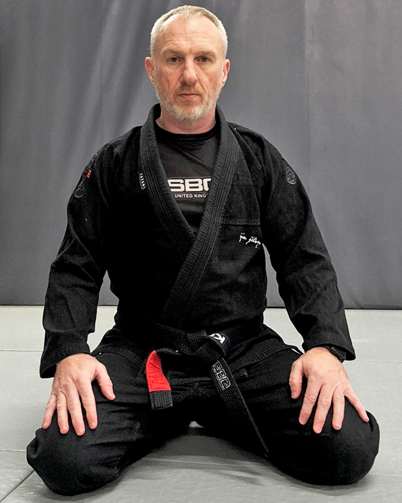 Simon Kenny kneeling on the mats in his black gi and black belt.